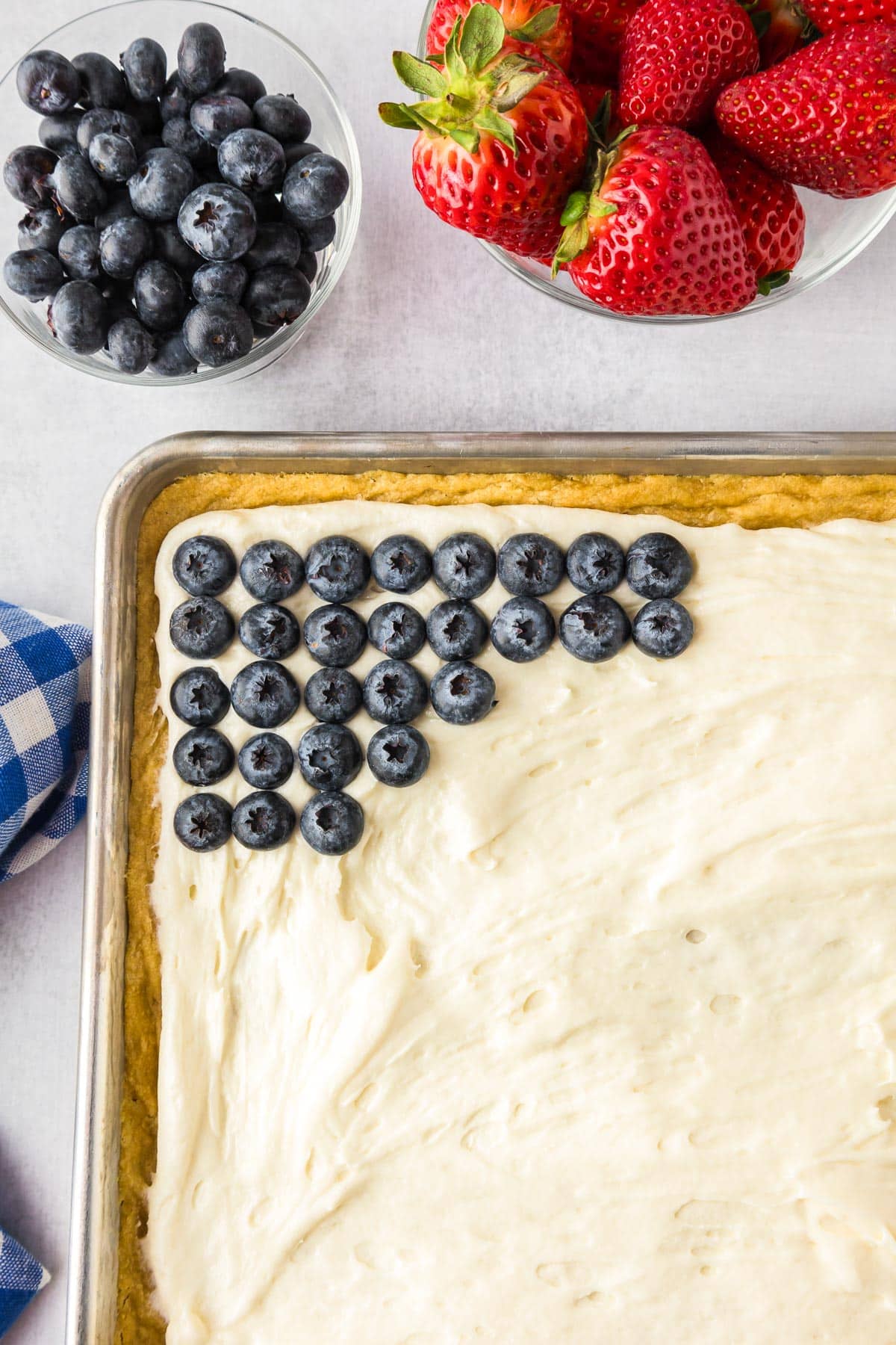 A rectangular baking pan with cream cheese frosting focused on the corner using blueberries to make the blue rectangle of the flag fruit pizza.