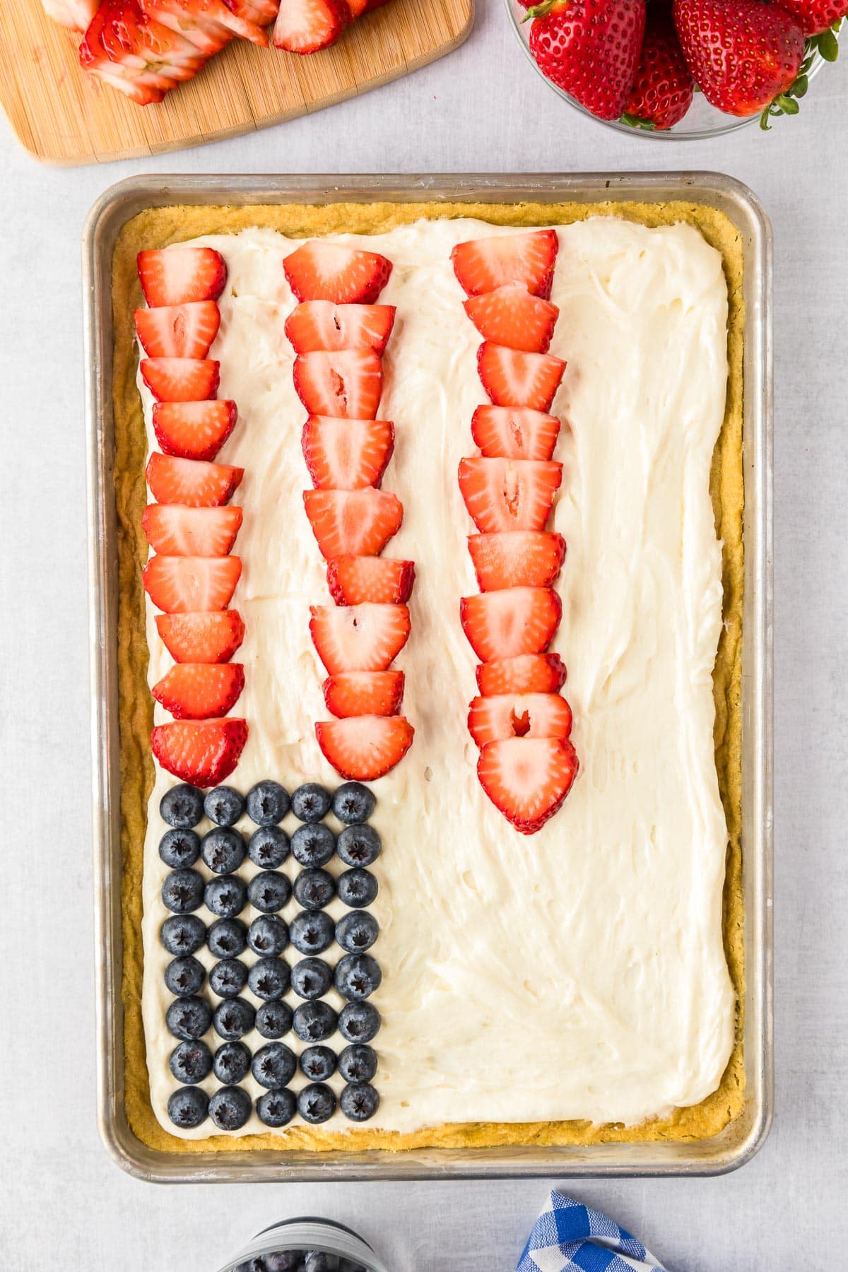 Laying pieces of strawberries for stripes on a large pan of sugar cookie dough to make a US flag with blueberries in a rectangle on the cookies.