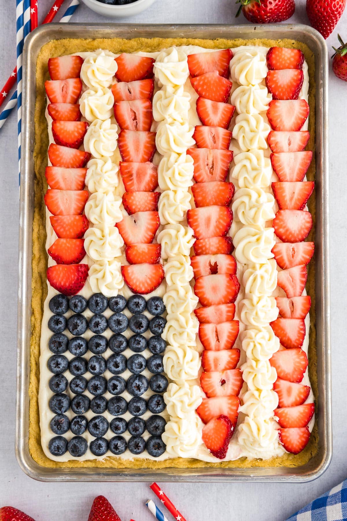 A large flag fruit pizza cookie resembling the American flag with rows of sliced strawberries, blueberries, and dollops of white frosting on a baking pan.