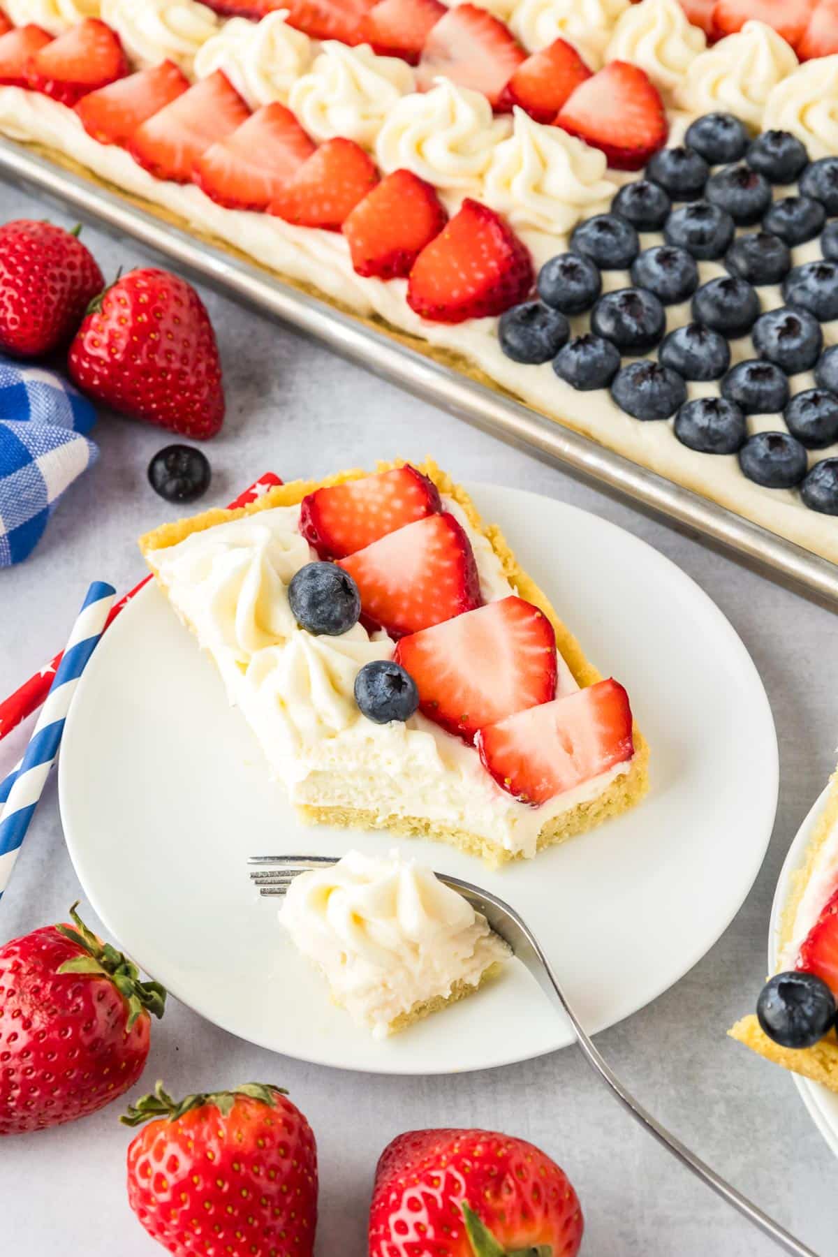 A slice of flag fruit pizza cookie being cut on a plate with a fork with a pan with flag fruit pizza in the background.