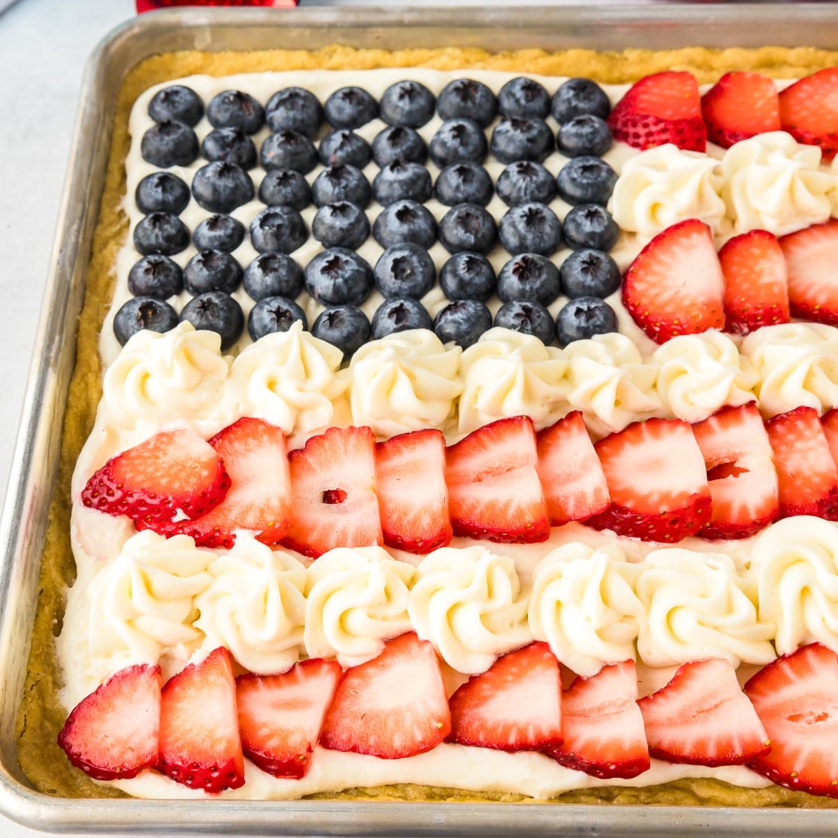 A flag fruit pizza decorated like a USA flag in a cookie pan with frosting and fruit decorations.