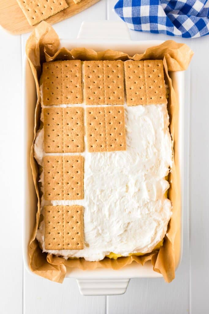 A baking dish with a layer of graham crackers being added to a thick layer of white marshmallow cream to make frozen lemon s'mores.