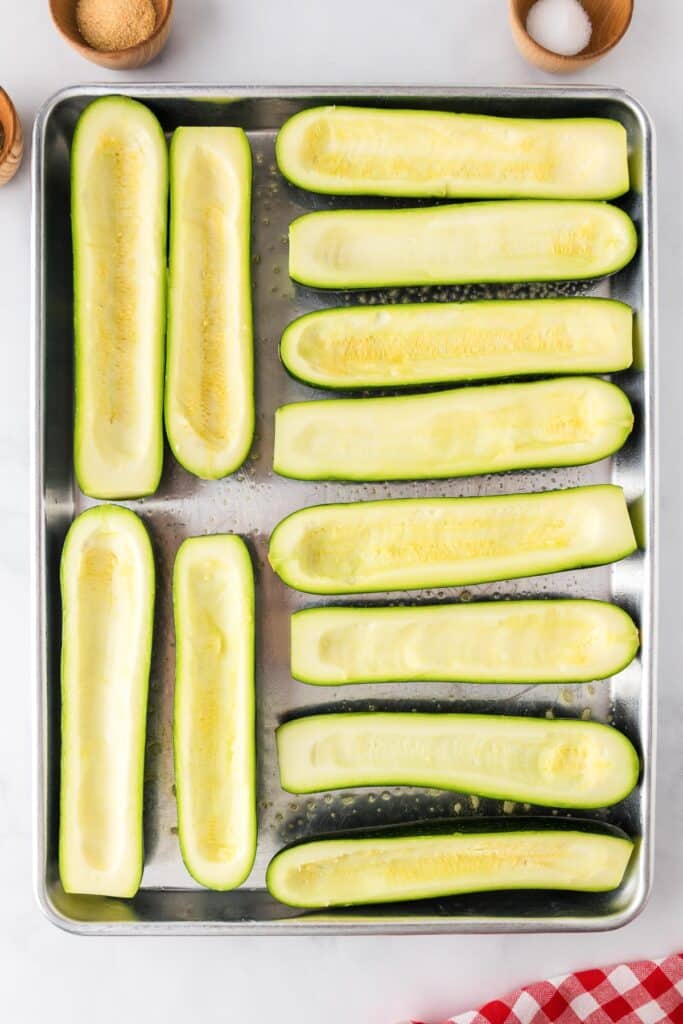 A baking sheet with ten halved and hollowed-out zucchini pieces on a counter.