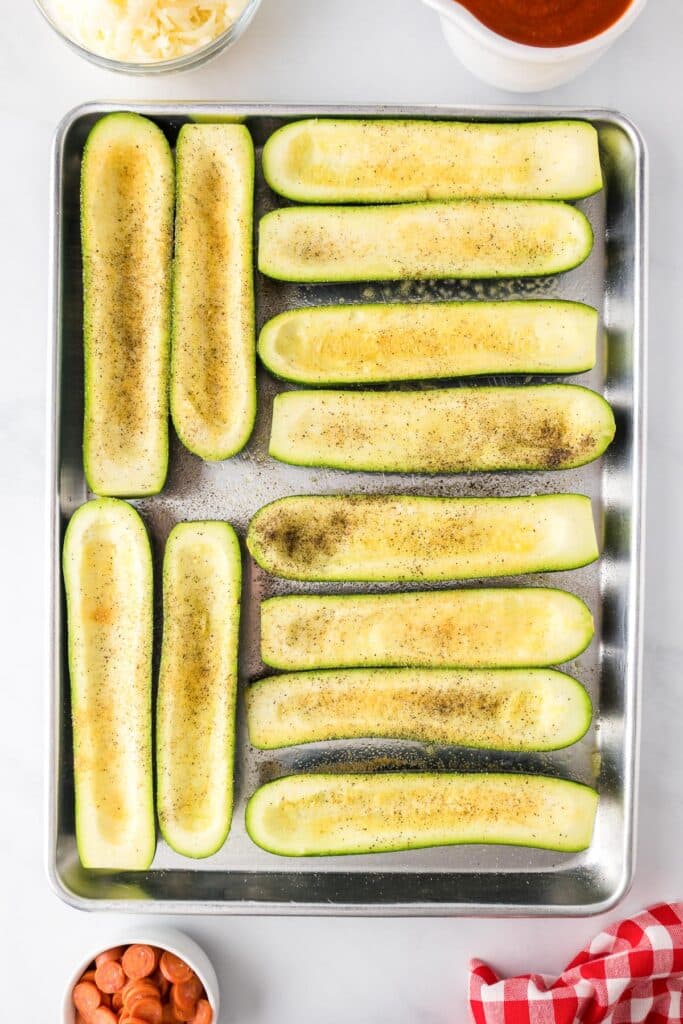 A baking tray with 10 halved zucchinis that have been hollowed out and seasoned.