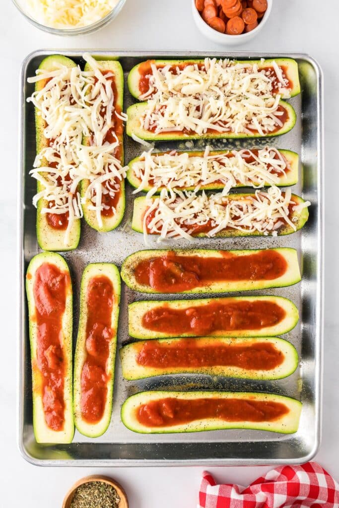 A baking tray with zucchini halves topped with marinara, and half topped with shredded cheese to make zucchini pizza boats.