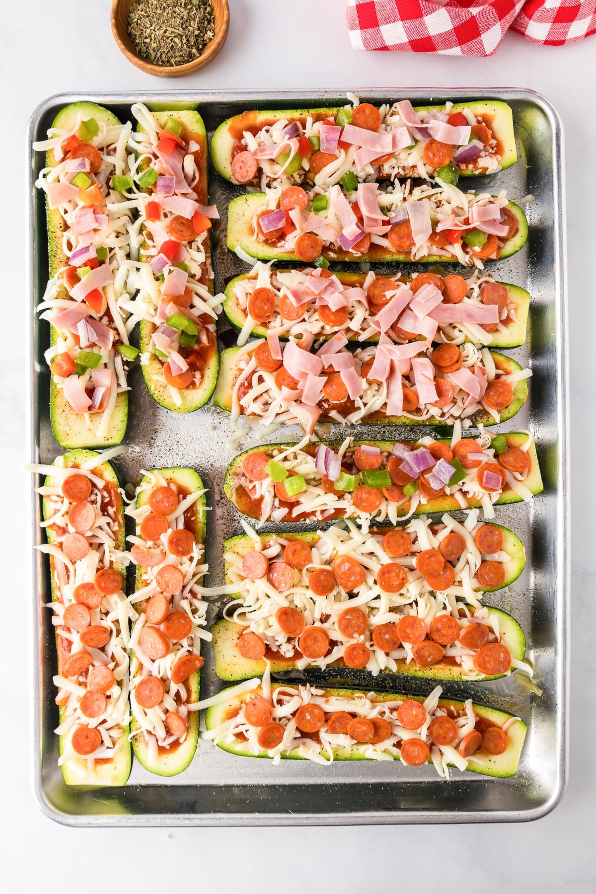 A baking tray filled with pizza zucchini boats topped with mini pepperoni, ham and diced vegetables before cooking.
