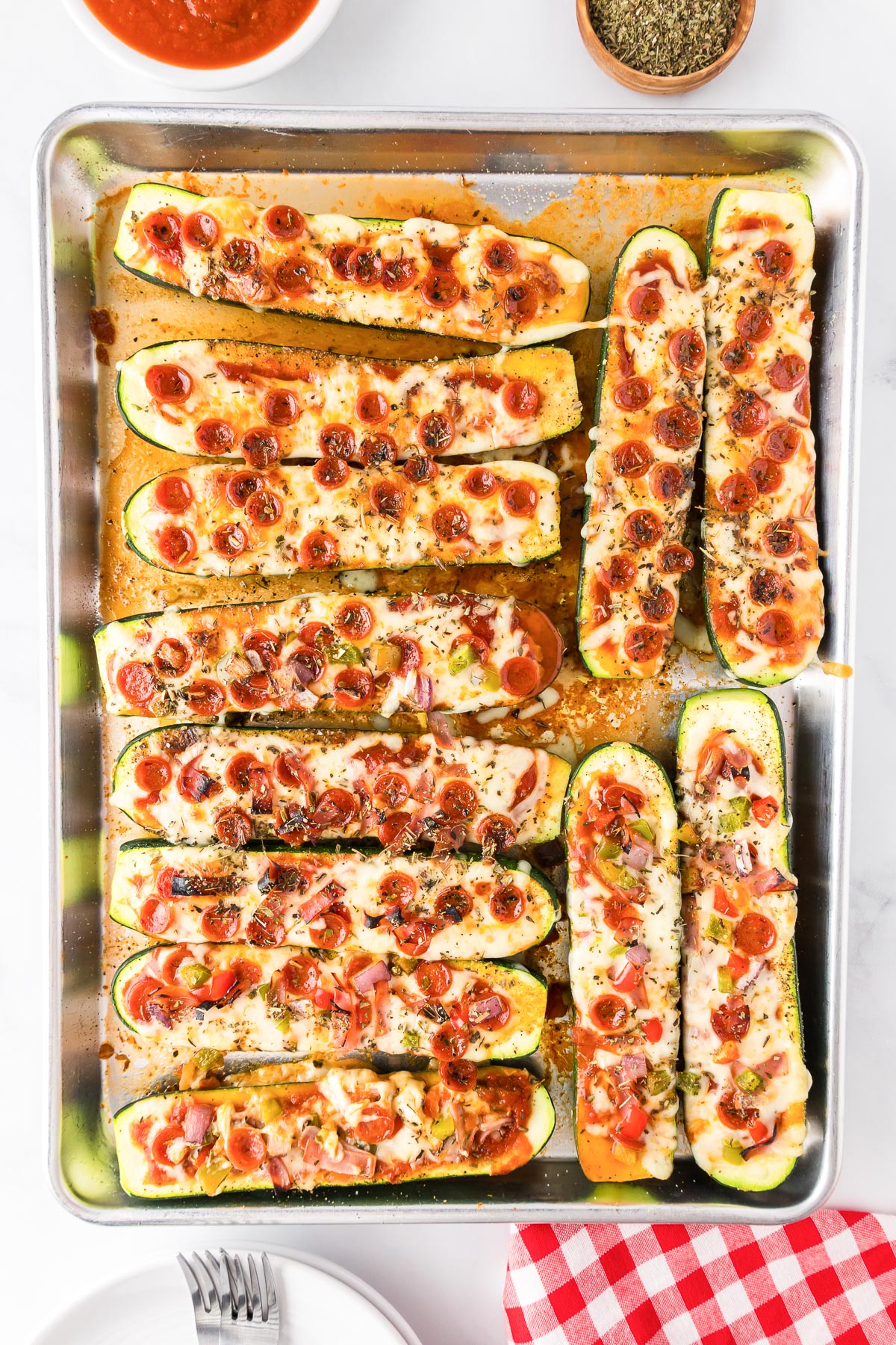 A large baking sheet filled with zucchini pizza boats topped with cheese, pepperoni, diced ham and other vegetable toppings.