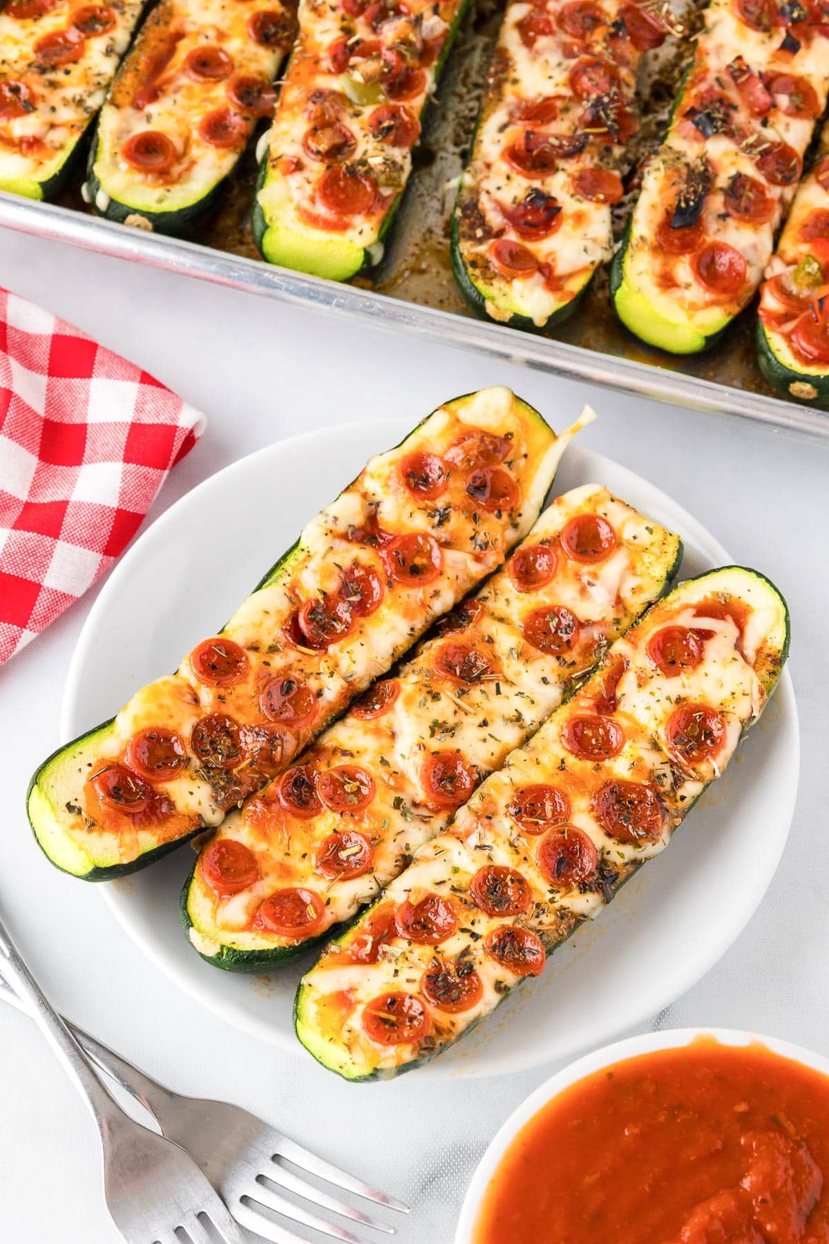 Three zucchini pizza boats topped with melted cheese and pepperoni on a plate with forks, a bowl of marinara and a pan full of more zucchini pizza boats in the background.