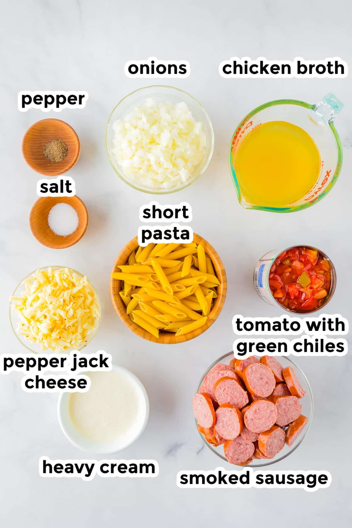 Overhead view of ingredients for cheesy smoked sausage pasta in bowls and measuring cups, labeled as smoked sausage, short pasta, chicken broth, onions, heavy cream, pepper jack cheese, tomato with green chiles, salt, and pepper.