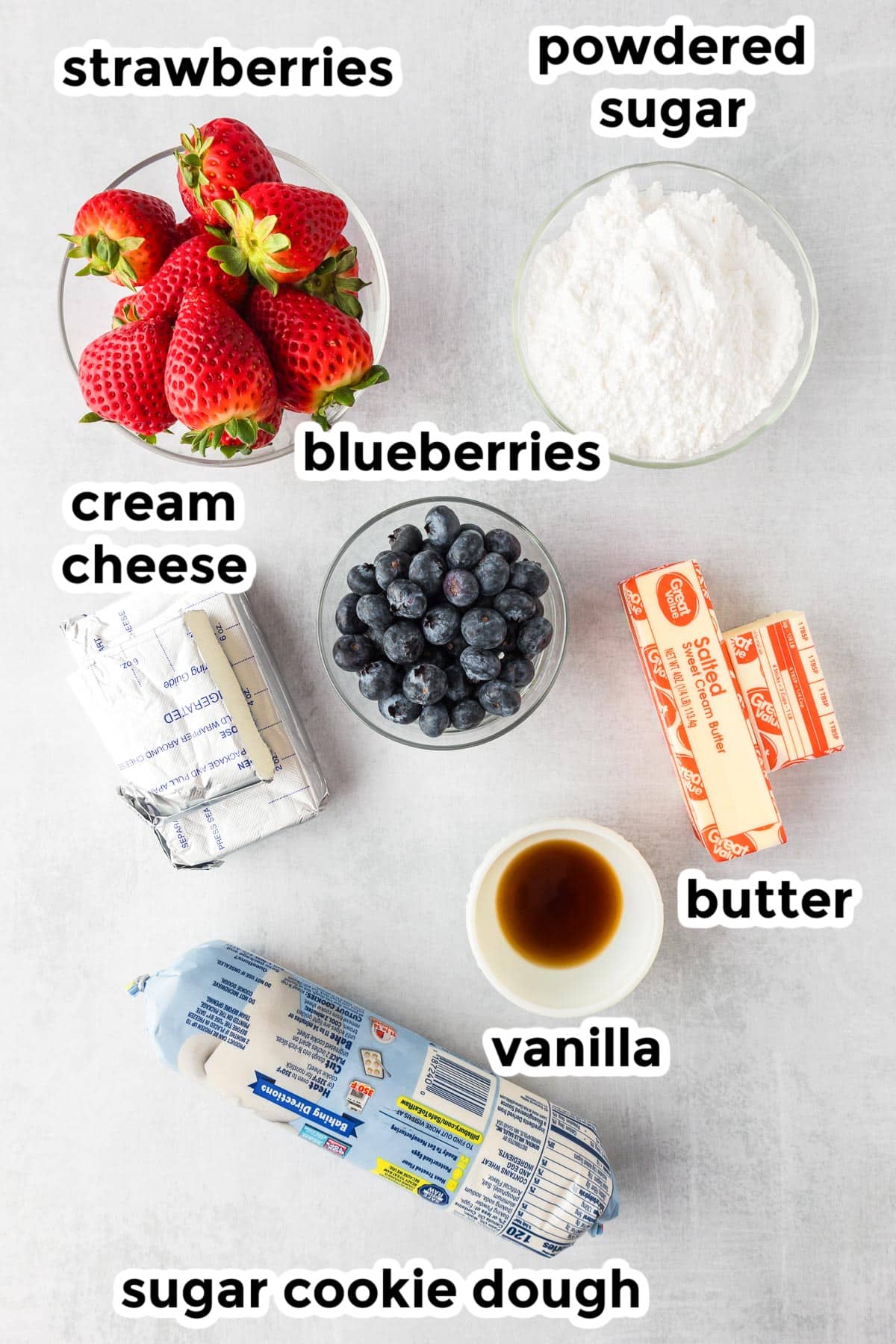 Ingredients for flag fruit pizza including strawberries, powdered sugar, cream cheese, blueberries, butter, vanilla extract, and a roll of sugar cookie dough on a counter with text labels.