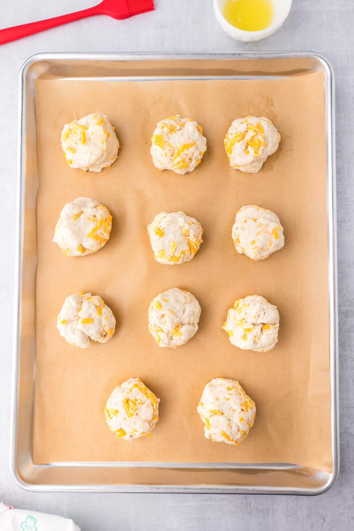 A baking sheet with 12 unbaked garlic cheese biscuits on parchment paper. A small bowl of melted butter and a red pastry brush are in the top left corner.