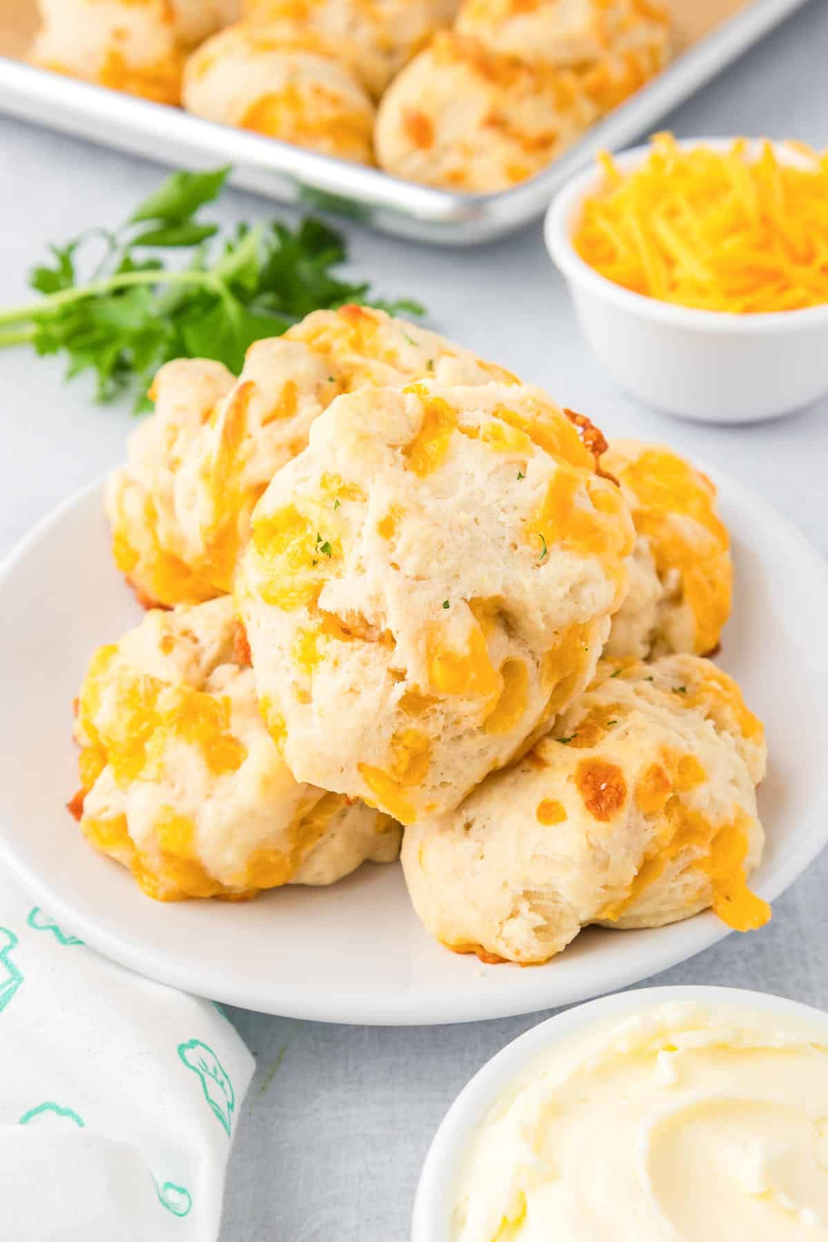 A plate of garlic cheese drop biscuits with more biscuits on a pan, cheese in a bowl, butter and a napkin around the biscuits.