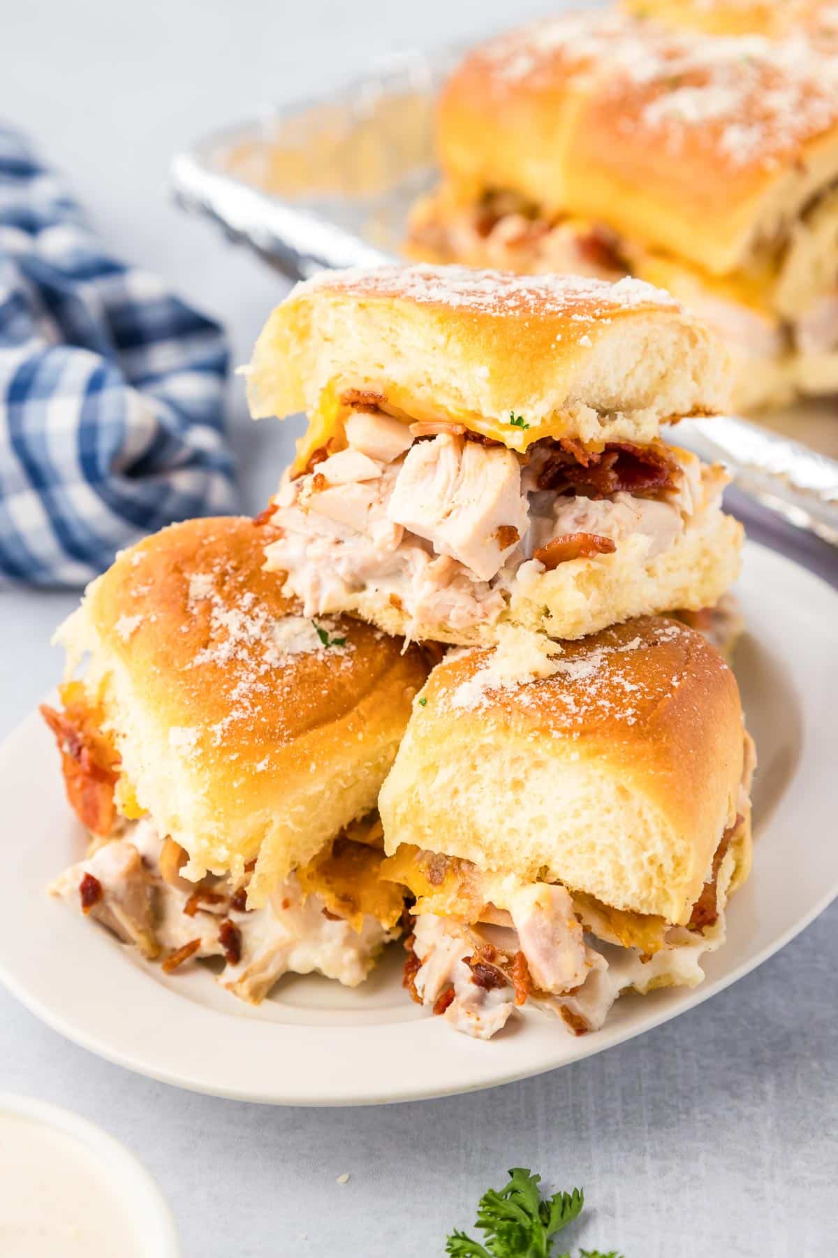 Three chicken bacon ranch sliders stacked on a serving plate with more in the background on a pan.