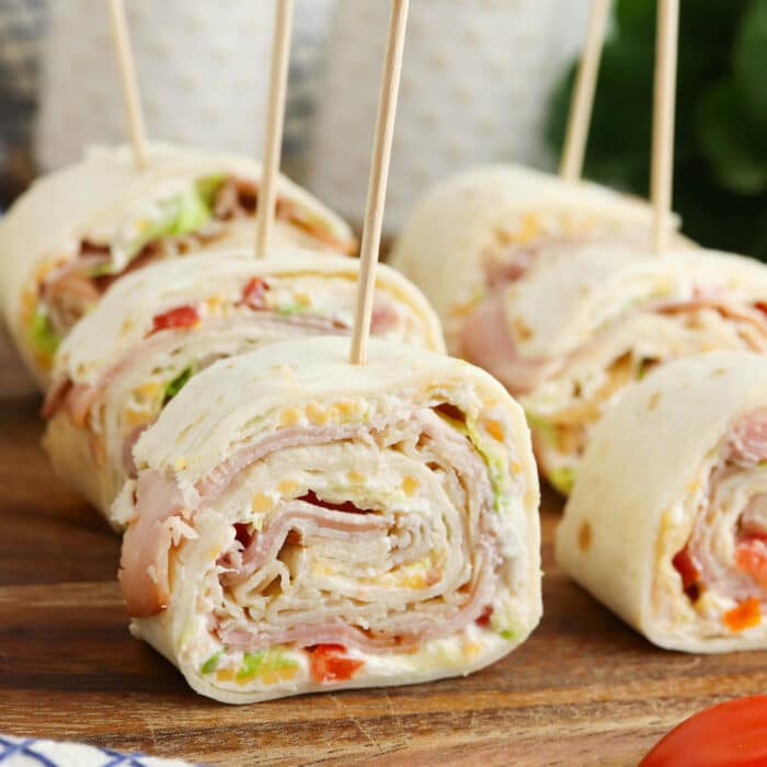 Four ham and turkey pinwheel sandwiches with toothpicks on a wooden cutting board from the side.