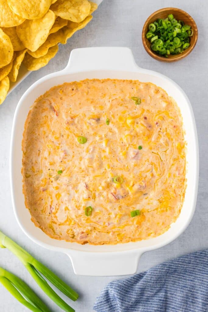 A square white dish filled with cheesy corn dip with Rotel with green onions and tortilla chips nearby.