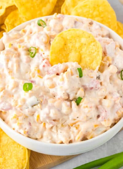 A bowl of creamy corn dip with Rotel next to tortilla chips with a chip in the dip.