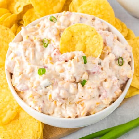 A bowl of creamy corn dip with Rotel next to tortilla chips with a chip in the dip.