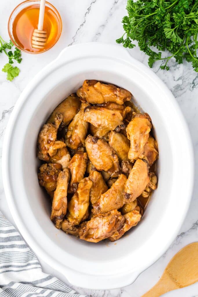 A white crockpot filled with cooked bbq chicken wings on a counter from overhead.