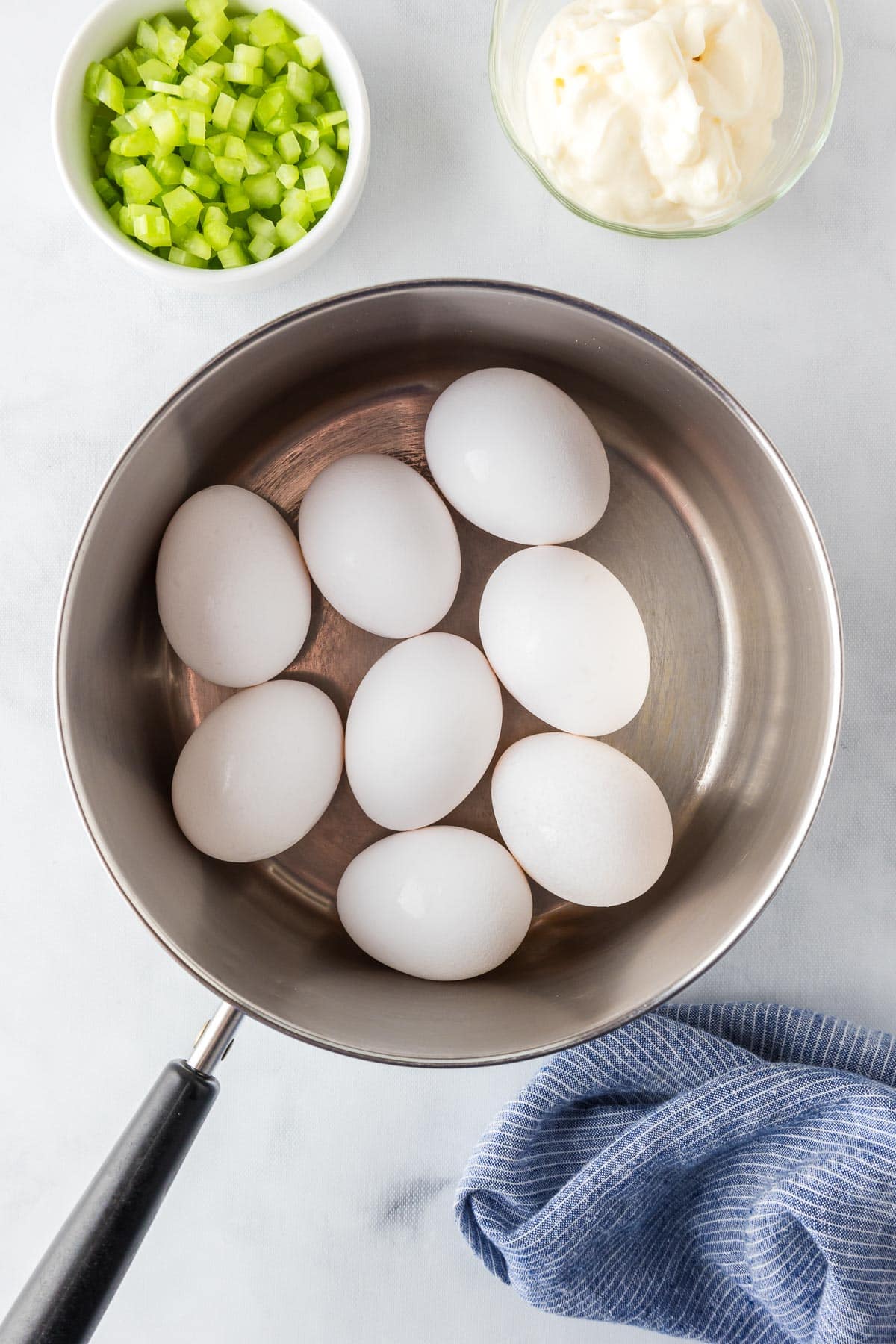 A pot containing white eggs in shells on a countertop with celery and mayonnaise nearby in bowls.