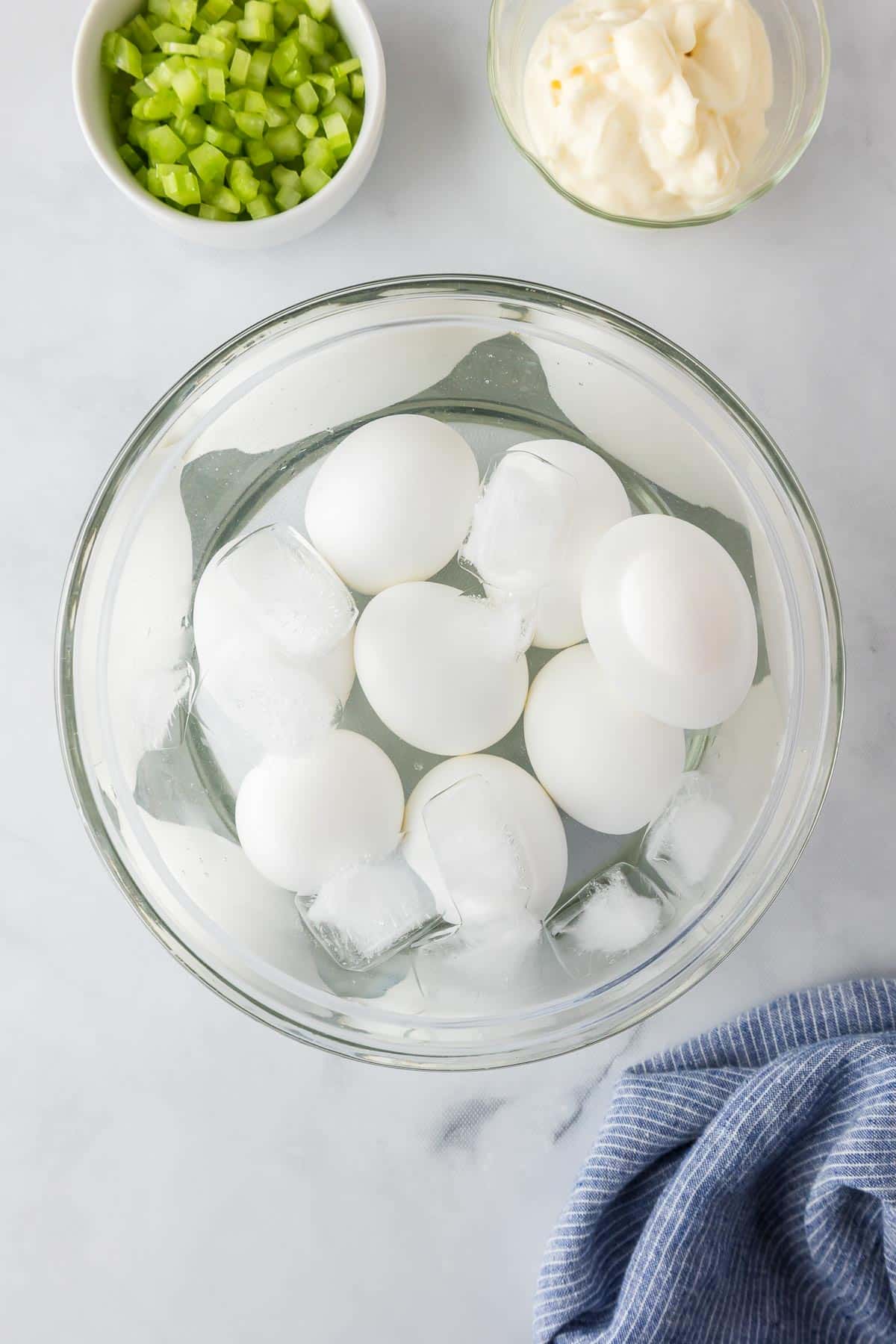 A bowl of hard-boiled eggs cooling in ice water, with bowls of chopped celery and mayonnaise nearby for egg salad.