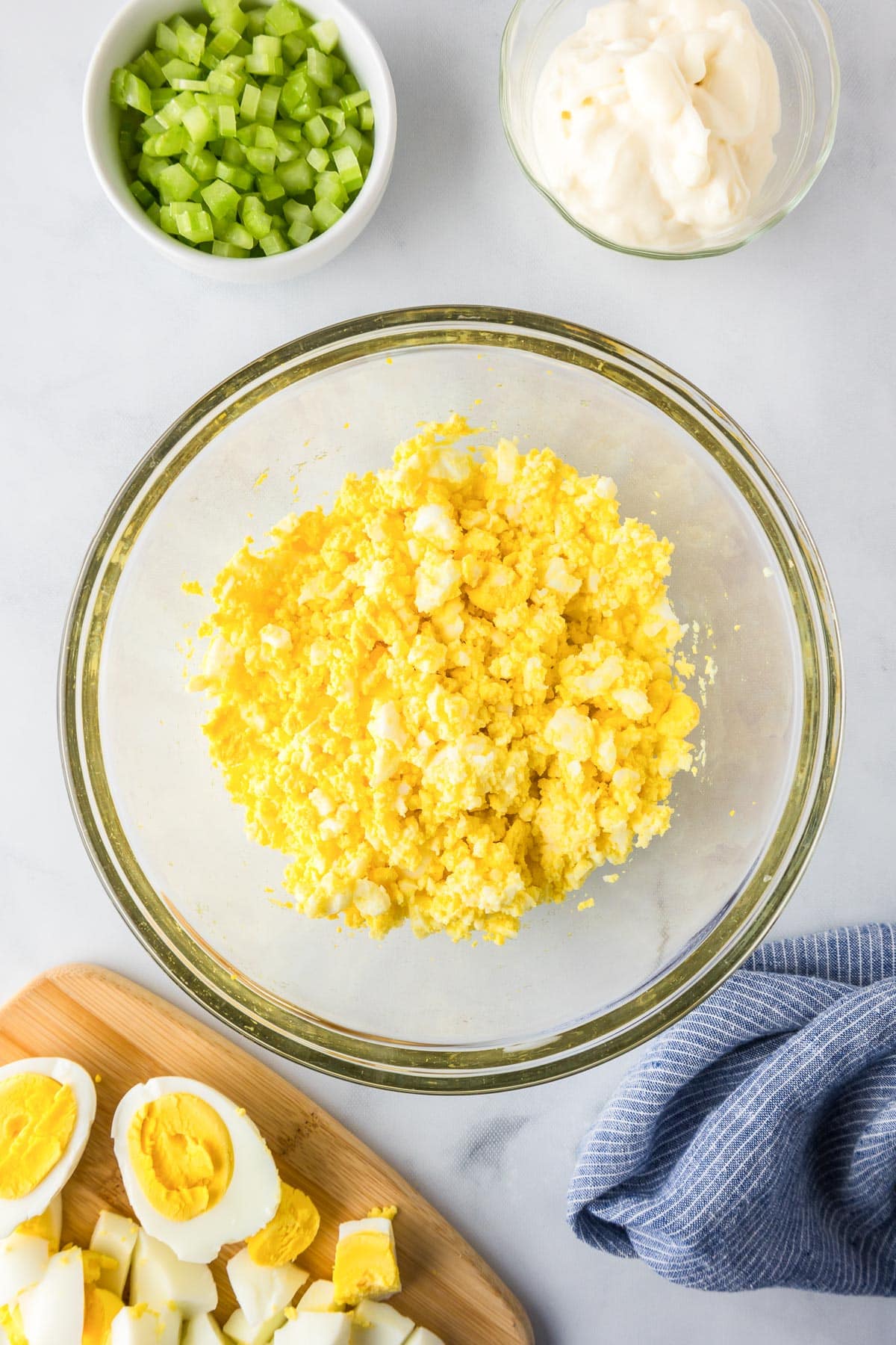A bowl of mashed egg yolks in a large bowl for egg salad with more hardboiled eggs, celery and mayonaise nearby in bowls.