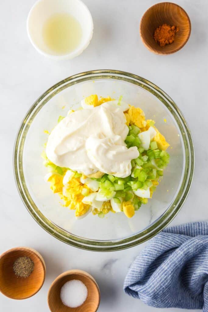 A glass bowl contains chopped hard-boiled eggs, celery, and mayonnaise with other spices and lemon juice in bowls nearby.