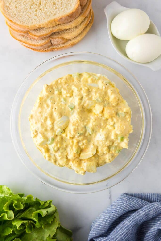 A clear bowl of egg salad surrounded by hard-boiled eggs, bread slices, a head of lettuce, and a blue-striped cloth on a marble surface.