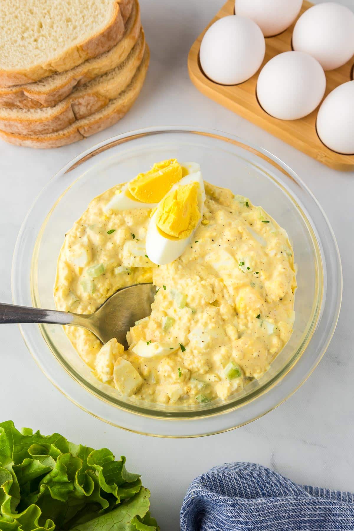 A bowl of egg salad with a spoon and bread and lettuce nearby to make sandwiches.