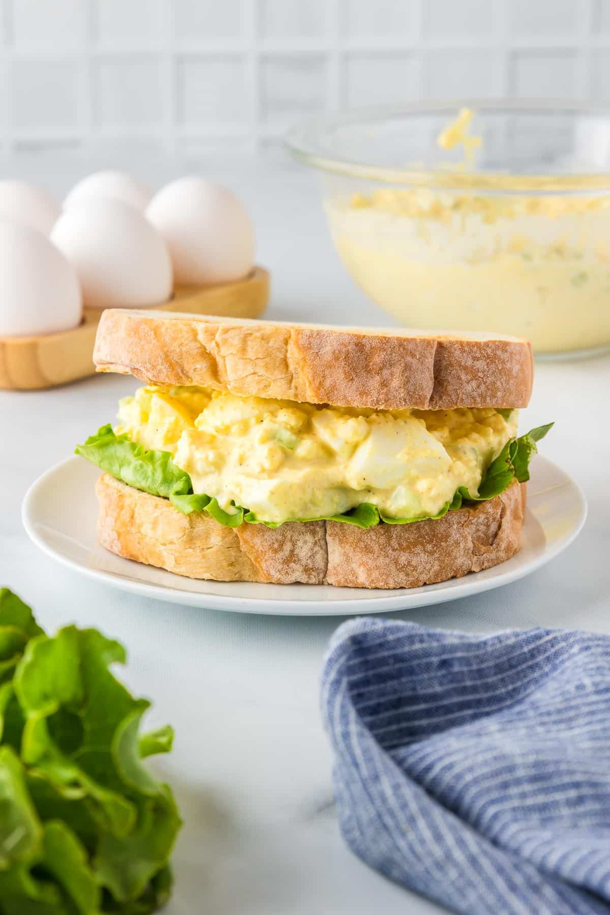 A plate holds an egg salad sandwich with lettuce on a plate with more ingredients in the background on the counter.