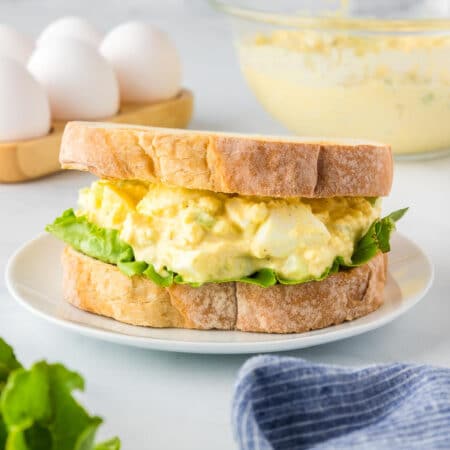 A sandwich with egg salad and lettuce on thick, toasted bread is on a white plate, with a bowl of egg salad, eggs in a carton, and a cloth napkin in the background.