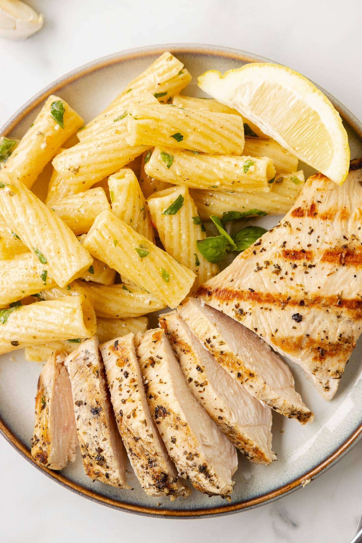 A plate with Italian grilled chicken breast with most of the chicken sliced and rigatoni pasta with herbs and a lemon wedge.