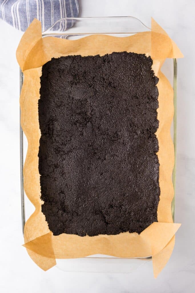 A rectangular baking dish lined with parchment paper and filled with a layer of dark, finely ground crumbs. A blue-striped cloth is partially visible in the top left corner.