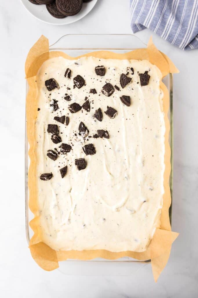 A rectangular dish lined with parchment paper full of cheesecake mixture with chopped Oreo pieces being sprinkled across the top.