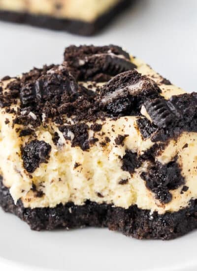 A close-up view of an Oreo cheesecake bar on a plate topped with Oreo cookie pieces with a bite missing.