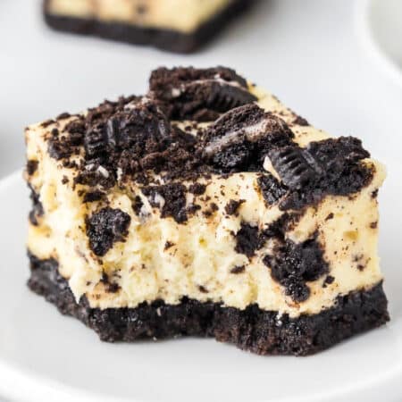 A close-up view of an Oreo cheesecake bar on a plate topped with Oreo cookie pieces with a bite missing.