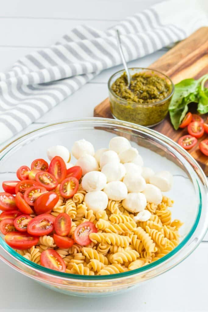 A glass bowl containing fusilli pasta, sliced cherry tomatoes, and mozzarella balls with pesto in a jar with a spoon in the background.