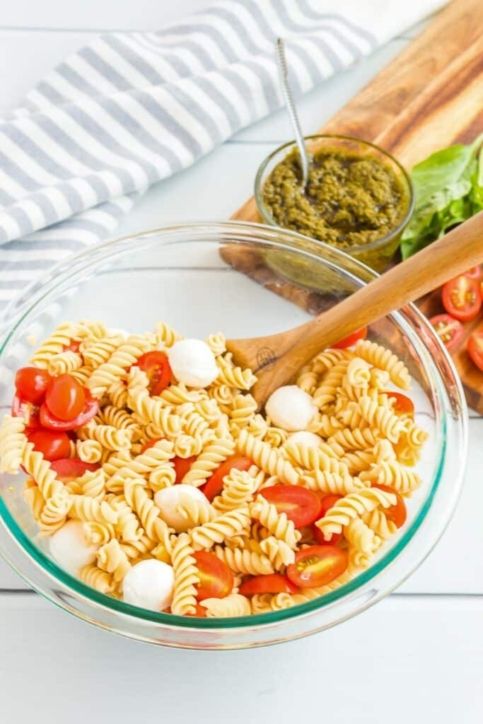 A bowl of pasta salad with rotini, cherry tomatoes, and mozzarella balls is mixed with a wooden spoon. A bowl of green pesto, fresh basil, halved cherry tomatoes, and a striped cloth are in the background.