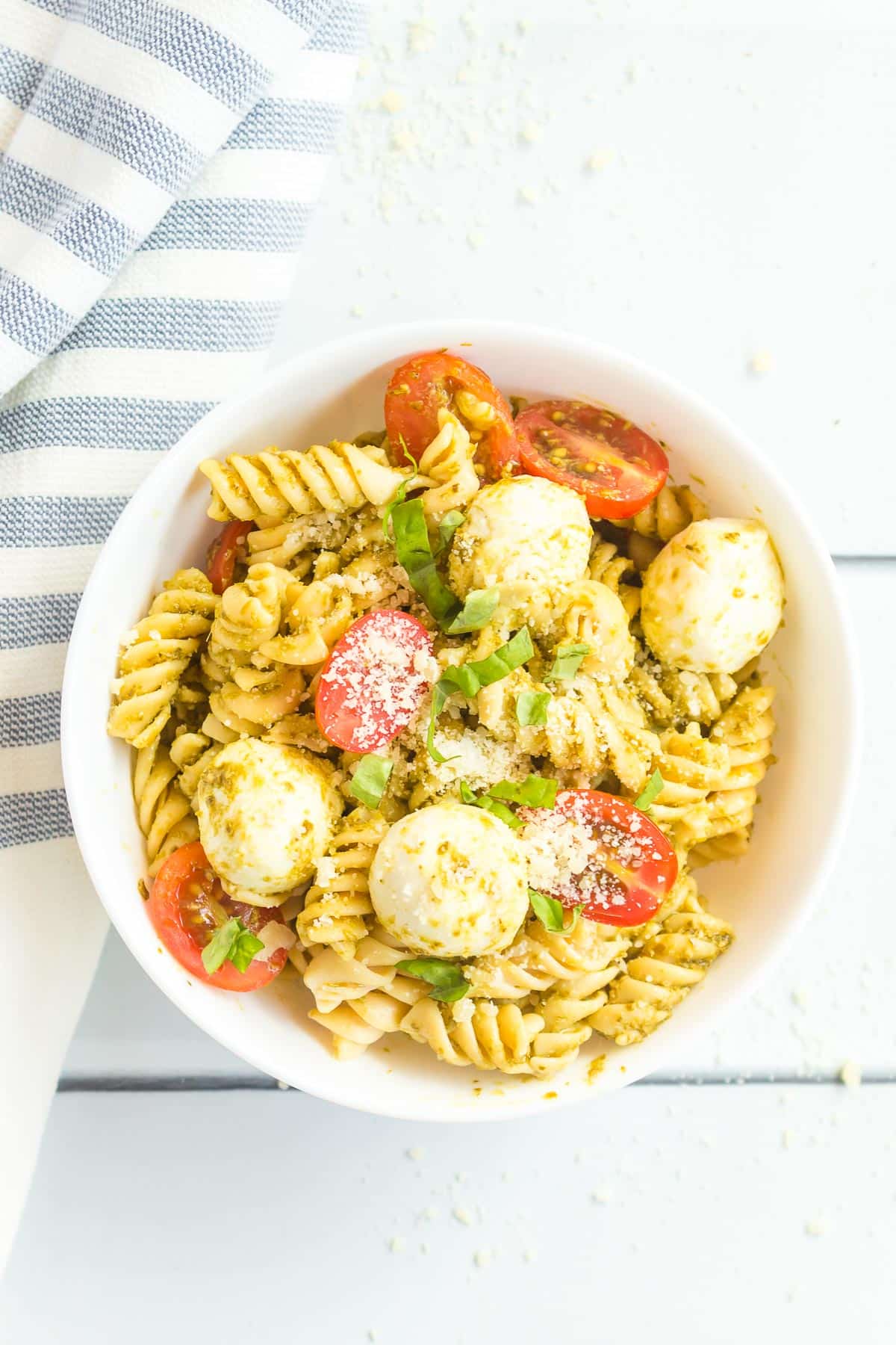 A bowl of pasta salad with cherry tomatoes, mozzarella balls, fresh basil, and sprinkled with grated parmesan cheese, next to a striped cloth on a white table.