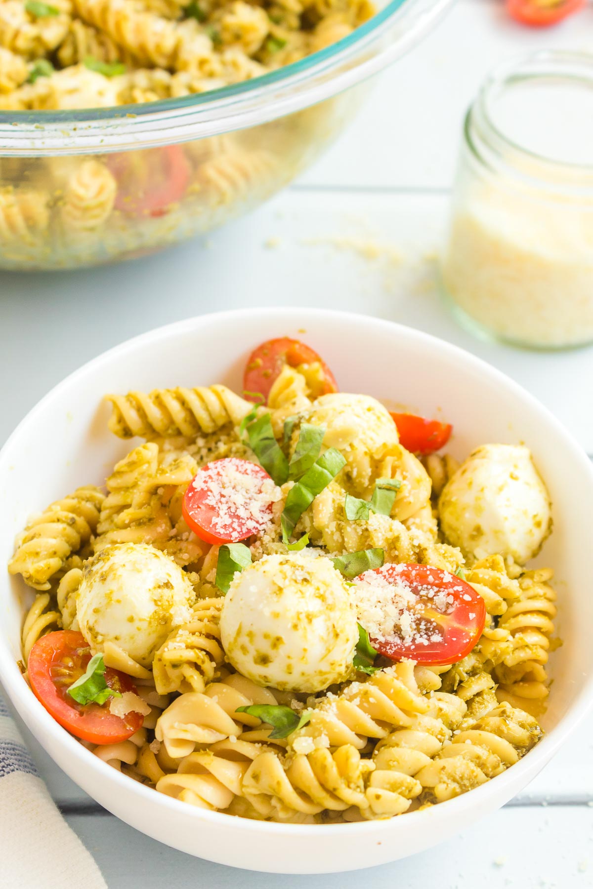 A bowl of pesto pasta salad at an angle topped with parmesan cheese with a larger bowl of pasta salad in the background/