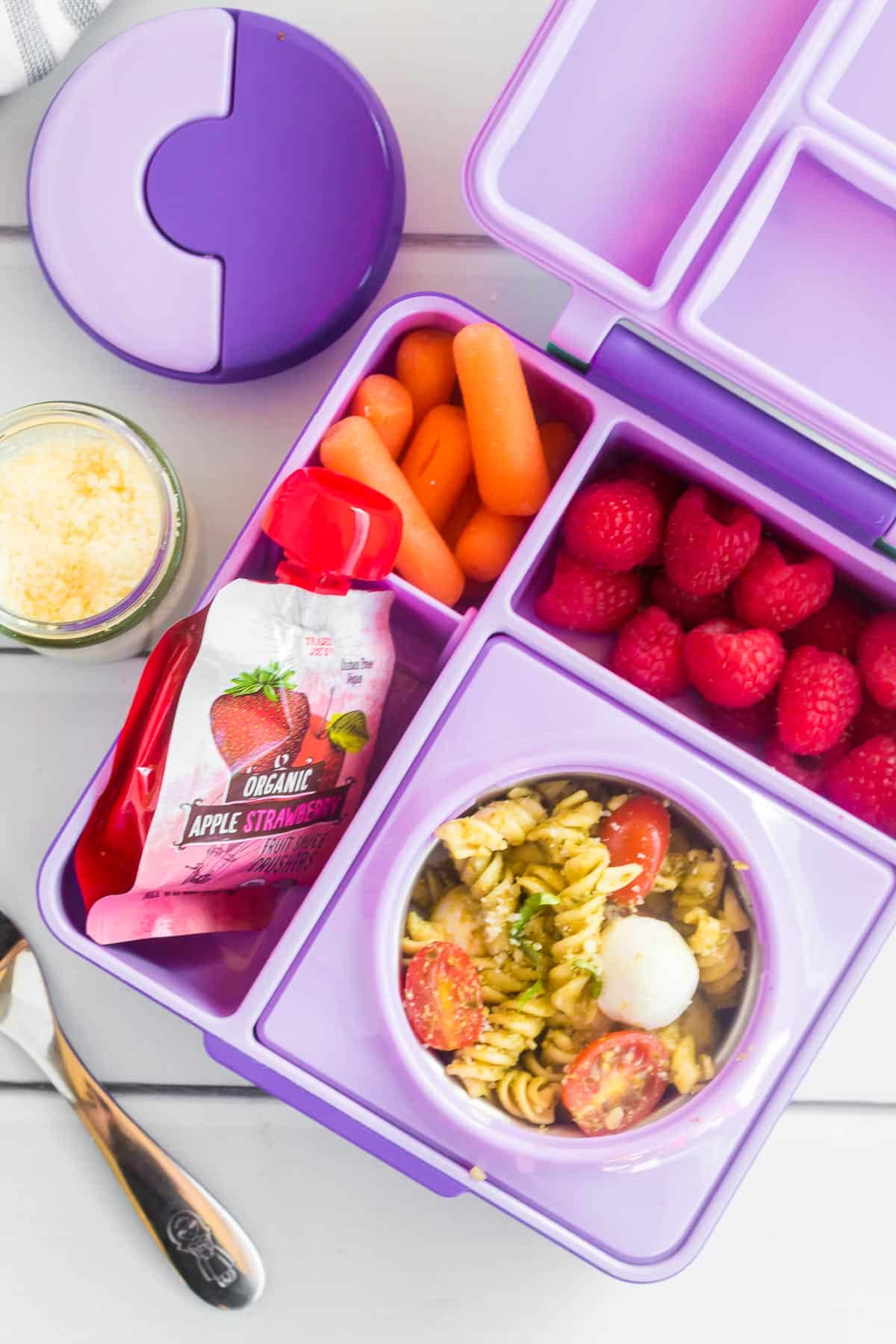 An open purple kids bento lunchbox full of pesto pasta salad, baby carrots, raspberries and an applesauce pouch from overhead.