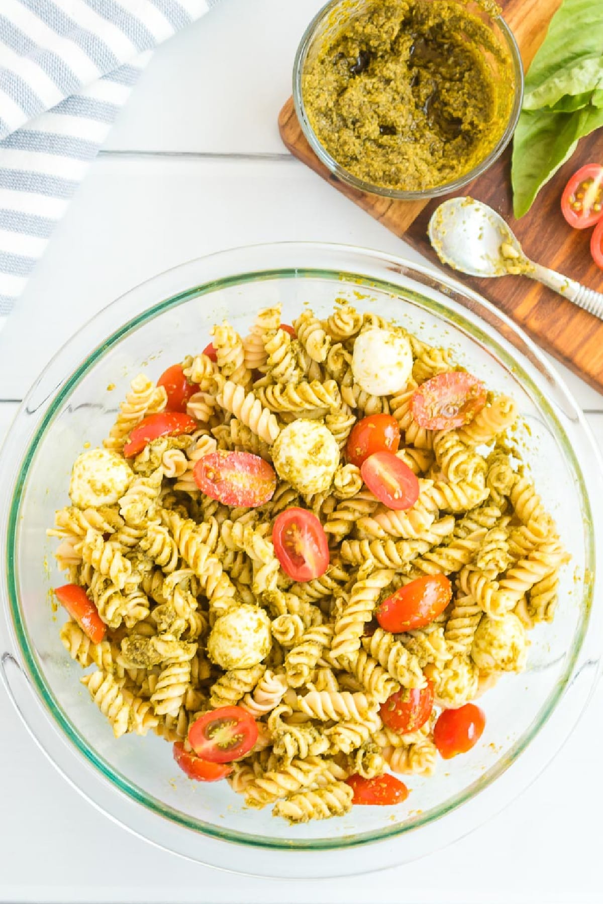 A bowl of pesto pasta salad with fusilli, cherry tomatoes, mozzarella balls, and pesto sauce. A spoon and a jar of pesto are on a wooden board nearby.