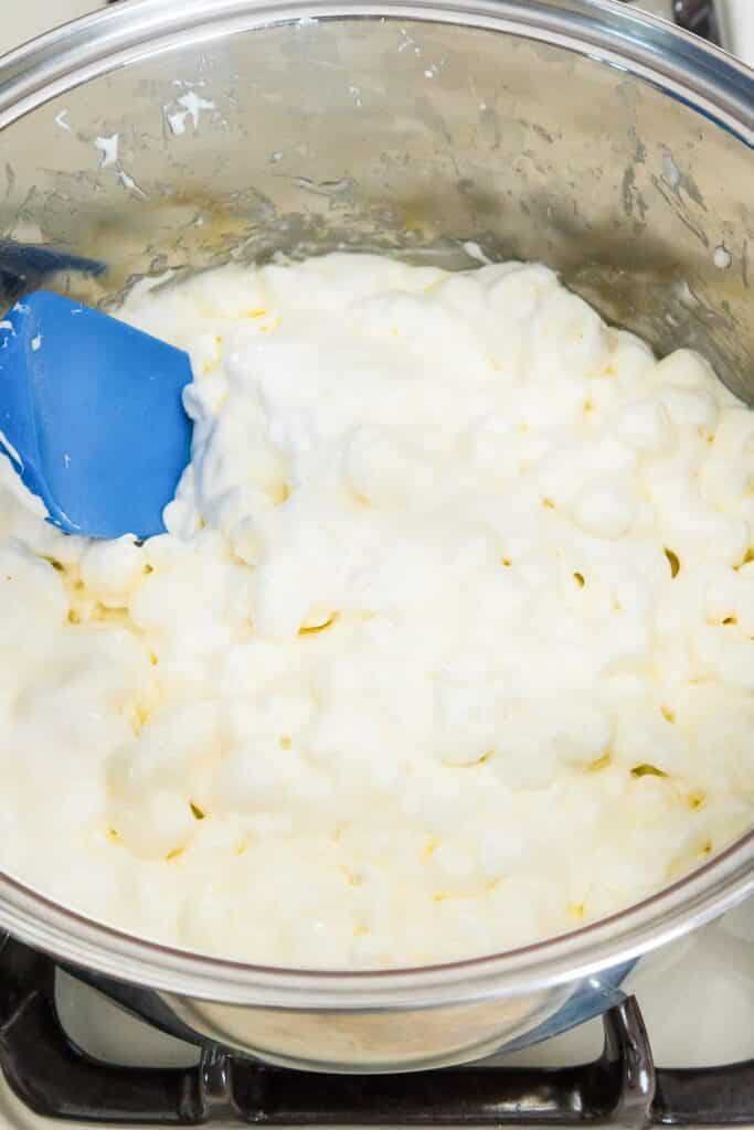 A close-up of a pot containing melted mini marshmallows being stirred with a blue spatula.