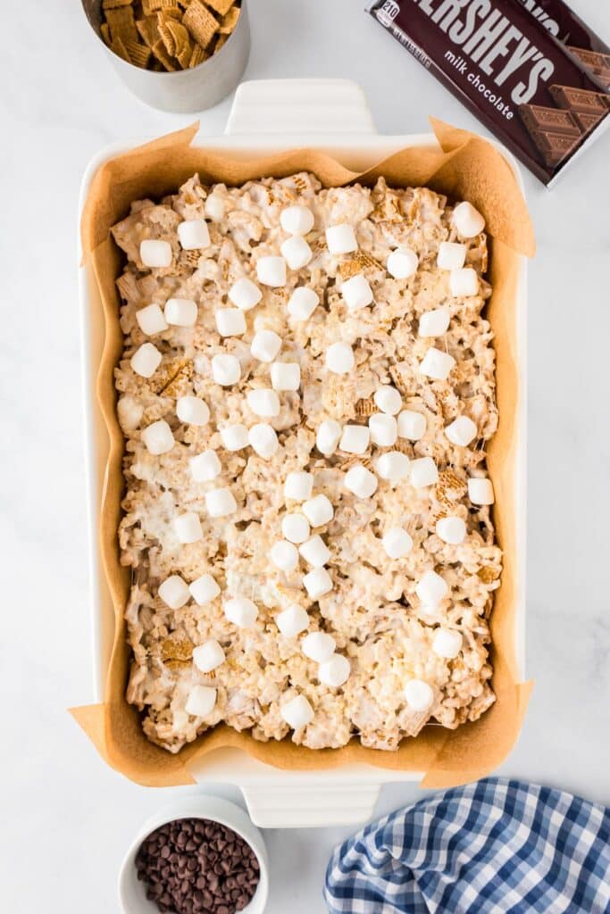 A tray of s'mores rice krispies treats topped with mini marshmallows and more chocolate pieces and Golden Grahams cereal pieces nearby.