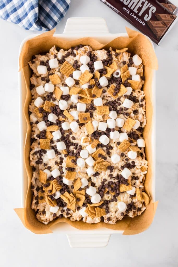 A baking dish with s'mores Rice Krispies treats topped with mini marshmallows, mini chocolate chips and Golden Grahams pieces on a counter.