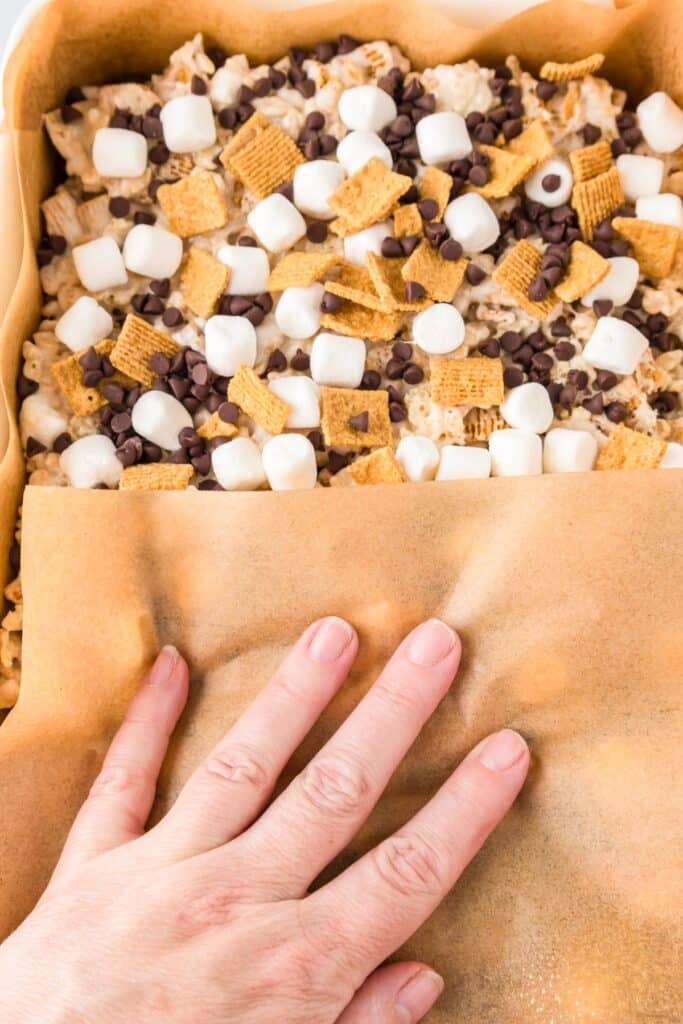 A hand gently presses down on a mixture of S'mores Rice Krispies treat bars with a piece of parchment paper.