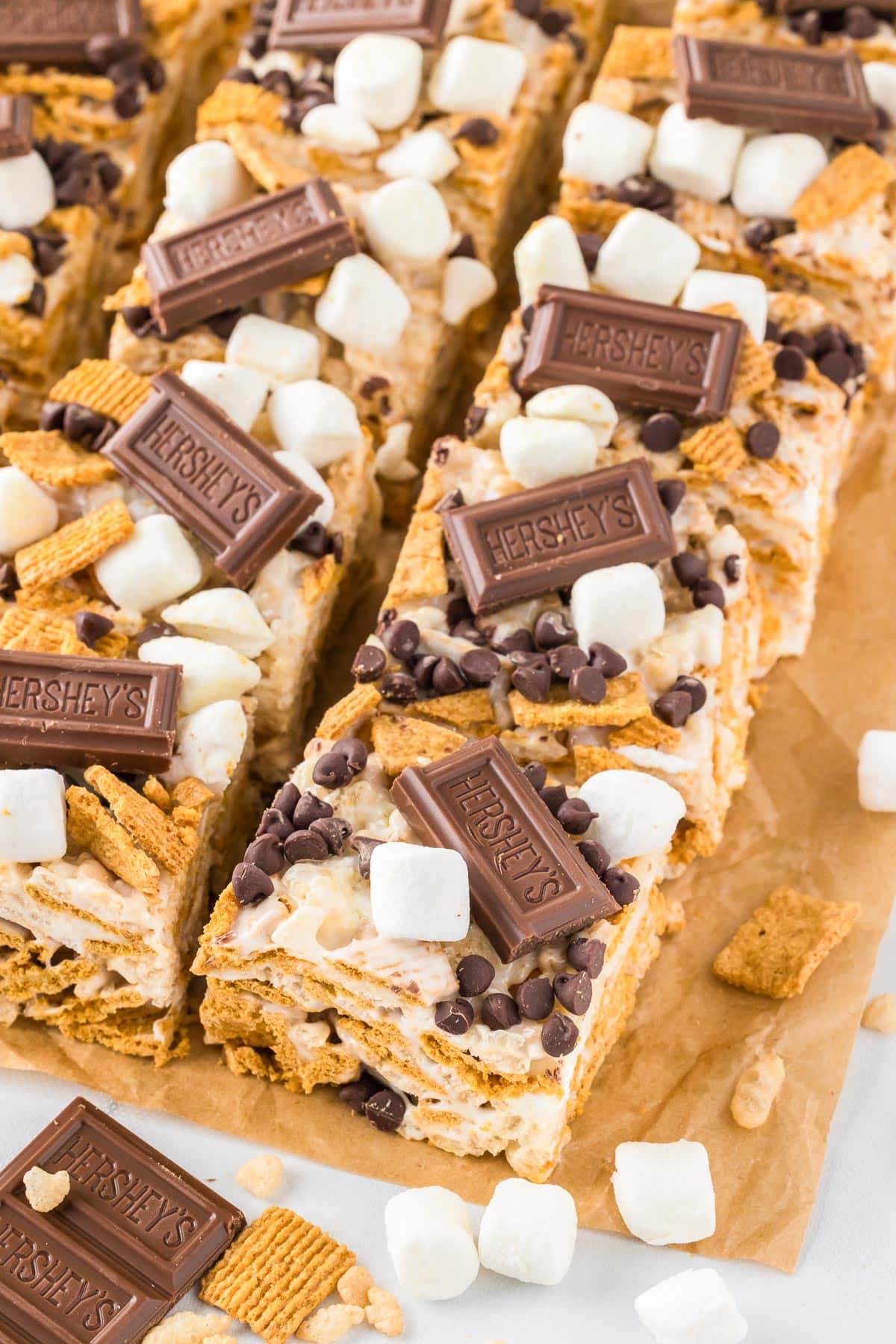 Close-up of s'mores Rice Krispies bars angled and sliced on a piece of parchment paper topped with pieces of Hershey's chocolate, mini marshmallows and mini chocolate chips.