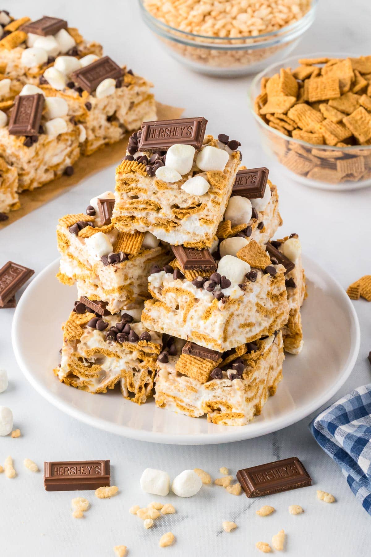 A plate of s'mores Rice Krispie treats bars stacked three high with more bars and bowls of cereal in the background.