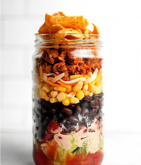 A layered taco salad in a mason jar featuring layers of meat, corn, black beans, salsa and corn chips on top.