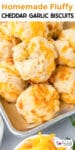 A tray of homemade fluffy cheddar garlic biscuits is shown close-up. The golden biscuits are topped with melted cheese and garnished with herbs.