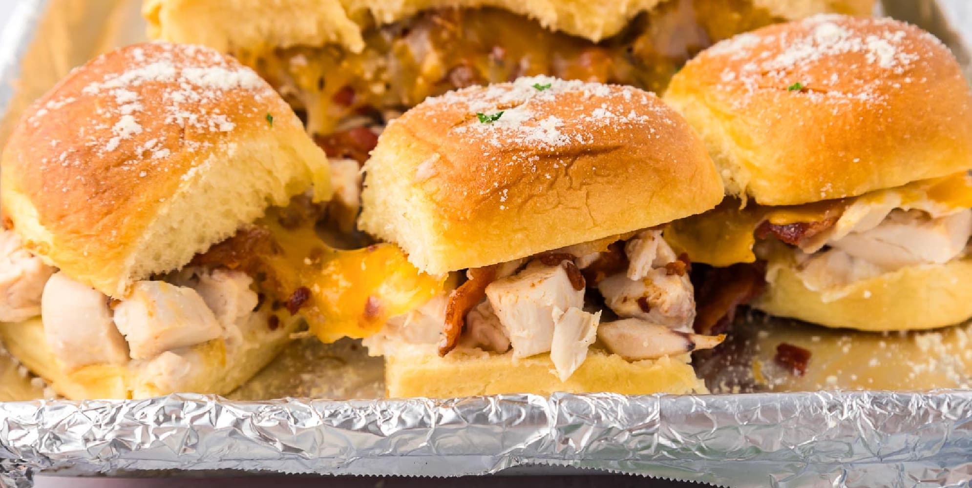 Three chicken bacon ranch sliders gooey on a aluminum foil lined pan from the side.